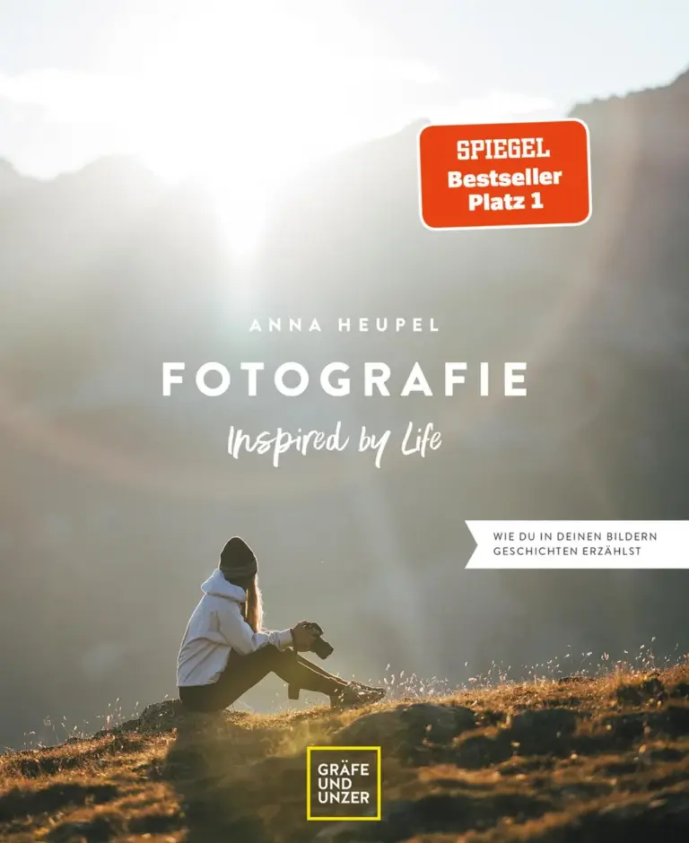Eine Frau mit Hut sitzt mit einer Kamera im Gras, Berge im Hintergrund, Titelbild eines Fotobuchs.