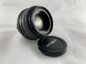 Ein Minolta-Kameraobjektiv mit dem Objektivdeckel daneben auf einer weißen Fläche.