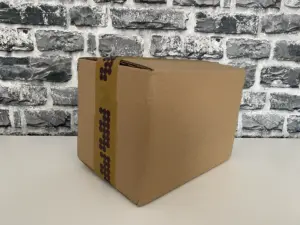 Ein geschlossener, mit braunem Klebeband versiegelter Karton steht auf einer weißen Fläche vor einer Backsteinmauer.