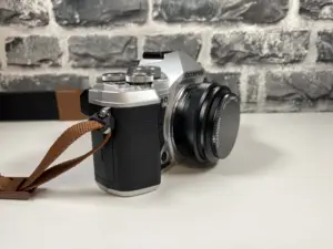 Eine silberne Olympus-Kamera mit einem braunen Riemen liegt auf einer weißen Fläche vor einer grauen Ziegelwand.