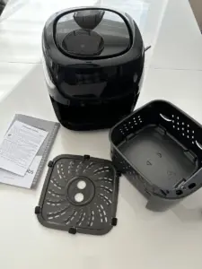 Eine schwarze Heißluftfritteuse mit Korb, Tablett und Bedienungsanleitung auf einer weißen Fläche.