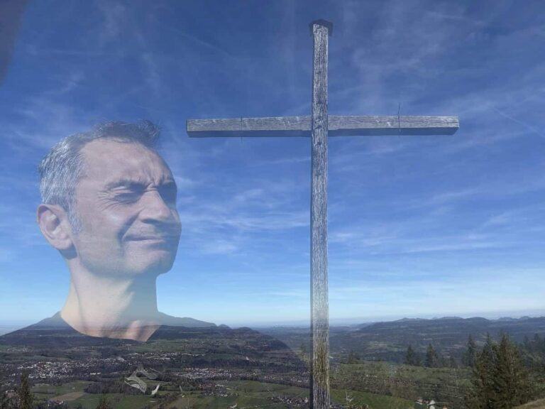 Das durchscheinende Gesicht eines Mannes erscheint am Himmel neben einem großen Holzkreuz auf einem Hügel mit einer malerischen Landschaft.