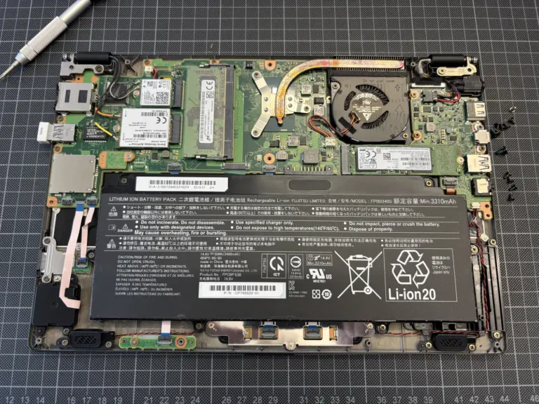 Geöffneter Laptop mit internen Komponenten: Akku, Lüfter, Hauptplatine, SSD und Schrauben auf einer schwarzen Arbeitsfläche mit Gittermuster.
