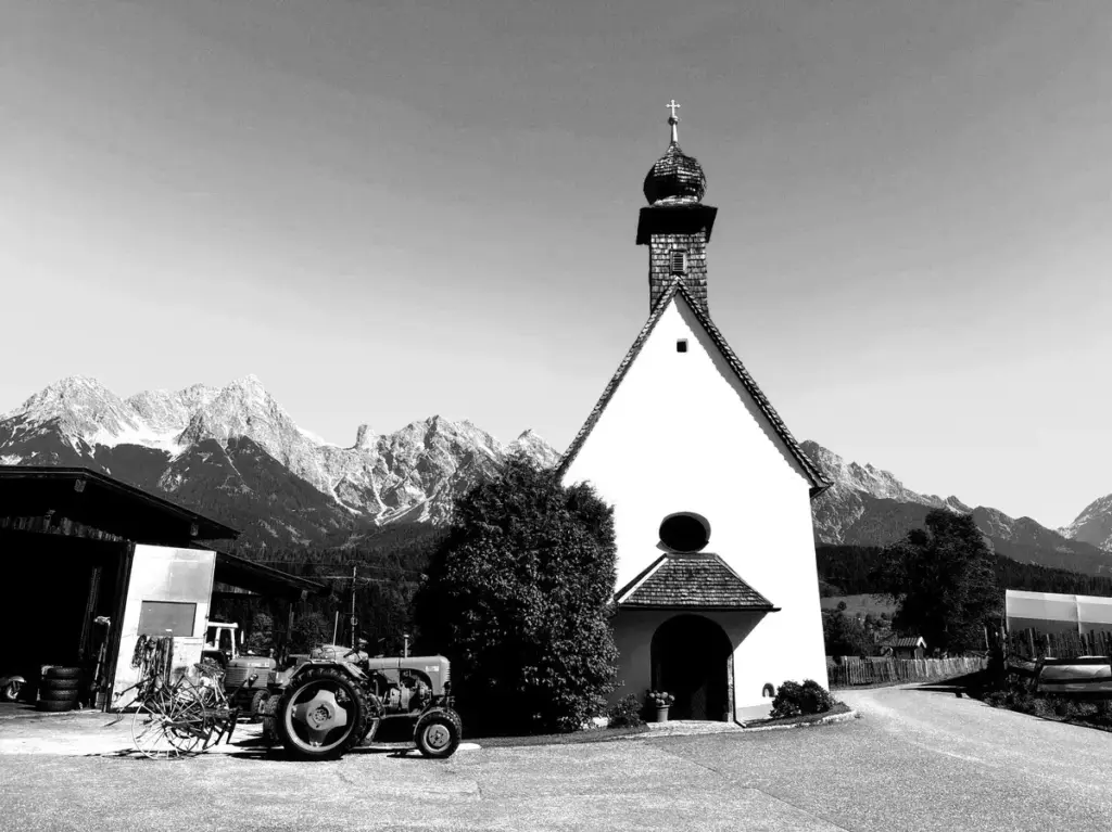 SW Aufnahme einer kleinen Kapelle, davor ein Oldtimer TRaktor, dahinter die Alpen